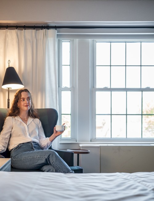 A woman relaxing in a cozy room, sitting in a high-backed chair, holding a cup, with natural light streaming through the window.