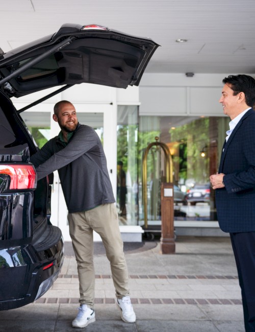 A man helps another load luggage into a car trunk while a third man, dressed in a suit, observes outside a hotel entrance.