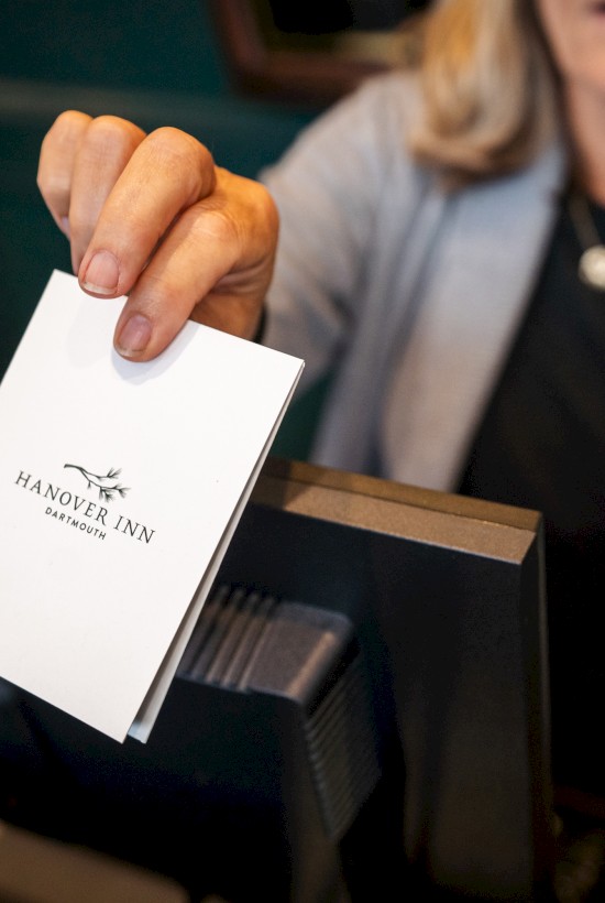 A woman hands a white card with the Hanover Inn Dartmouth logo at a hotel reception desk.