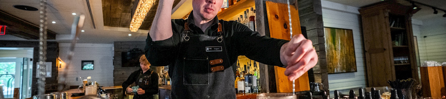 A bartender in a black apron is preparing a cocktail at a bar inside a cozy, warmly lit restaurant or bar.