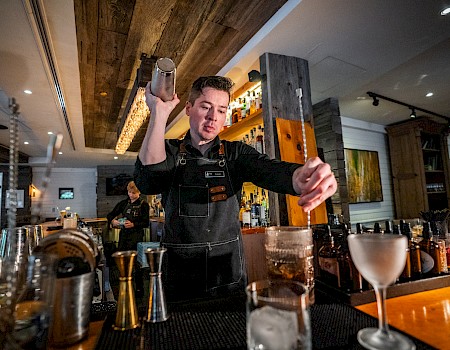 A bartender in a black apron is preparing a cocktail at a bar inside a cozy, warmly lit restaurant or bar.