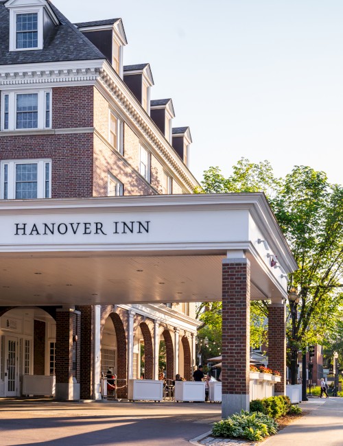 The Hanover Inn is a historic hotel with brick architecture, white accents, outdoor seating, and lush green trees.