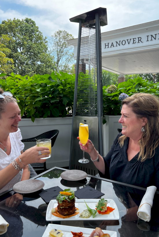 Two women enjoy drinks and food outdoors at a restaurant patio, celebrating together with smiles in front of a heater.