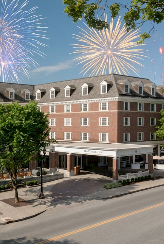 Fireworks light up the sky over a hotel named Hanover Inn on a clear day with green trees and outdoor seating in front.