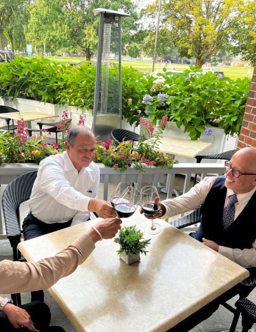 Three people clink glasses filled with red wine at an outdoor table amid lush greenery and colorful flowers.