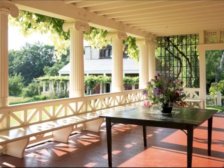 A porch with white columns, a wooden table with a floral arrangement, and lush greenery outside on a sunny day.