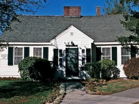 A cozy white house with black shutters, surrounded by trees and bushes, under a clear blue sky, exuding charm and tranquility.