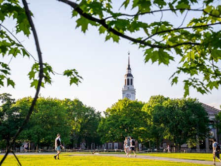 A park scene with students walking on grass, surrounded by green trees, and a tall clock tower in the background under a bright sky.