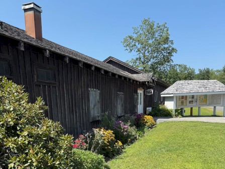 A rustic black wooden building with colorful flowers in front, accompanied by an information display case, under a clear blue sky.