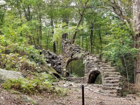 A stone archway remains of an old structure in a wooded area, with trees and greenery surrounding it and a small pathway in the foreground.