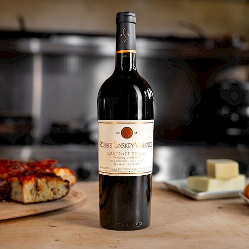 A bottle of Robert Sinskey Vineyards Cabernet Franc wine stands on a kitchen counter with pizza and cheese in the background.