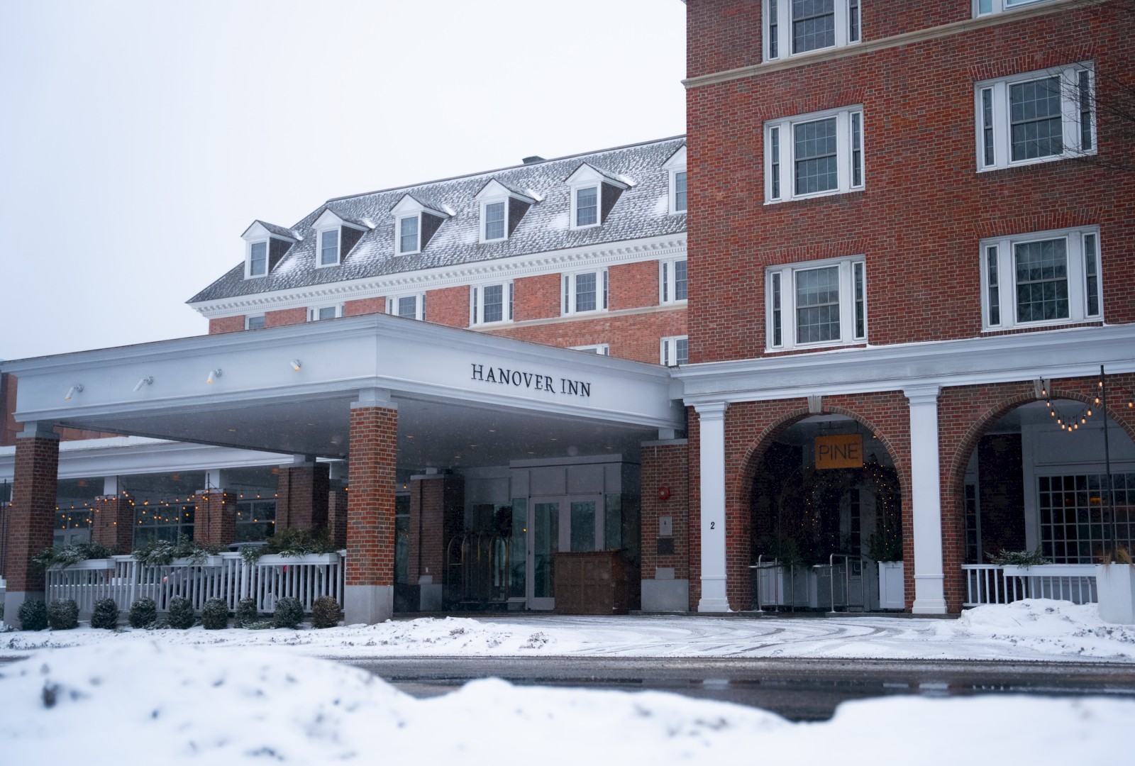 The image shows the exterior of Hannover Inn, a brick hotel building during snowy weather with a covered entrance and string lights.