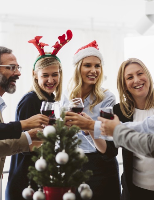 A diverse group of people celebrating Christmas, wearing festive hats, raising glasses in a cheerful toast, around a small decorated tree.