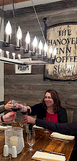 Three women are celebrating with glasses of red wine at a dinner table in a cozy, rustic restaurant called The Hanover Inn Coffee Shop.