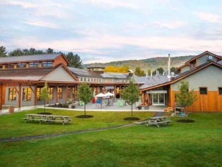 A large, modern building with wooden accents, outdoor seating, picnic tables, trees, and a grassy area under a partly cloudy sky.