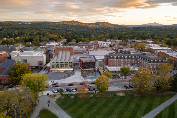 A scenic aerial view of a small town centerpiece with shops, trees, parking, and a park in the foreground, surrounded by hills at sunset.