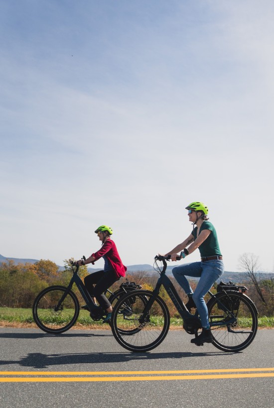 Two women ride e-bikes with helmets on a sunny, open road, surrounded by trees and hills in the distance.
