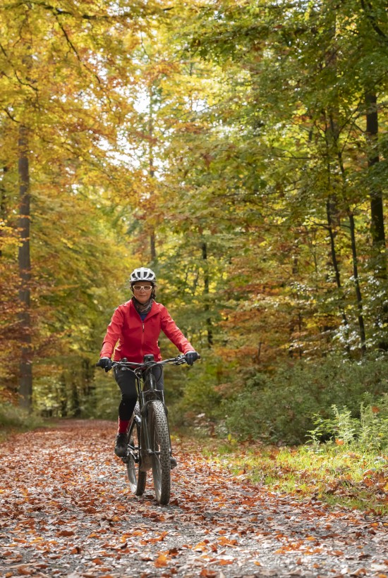 A woman in a red jacket rides a mountain bike on a leaf-covered trail through a colorful autumn forest.