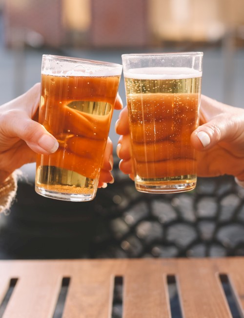 Two people are clinking glasses of beer outdoors, celebrating together on a sunny day.