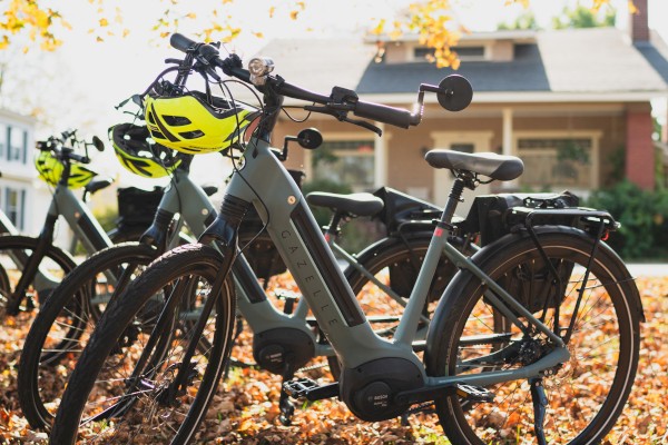 Electric bicycles parked outdoors on a fall day, with colorful leaves on the ground and houses in the background.