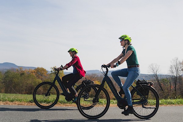Two women are riding e-bikes on a scenic road, wearing helmets with a backdrop of hills and a clear sky.