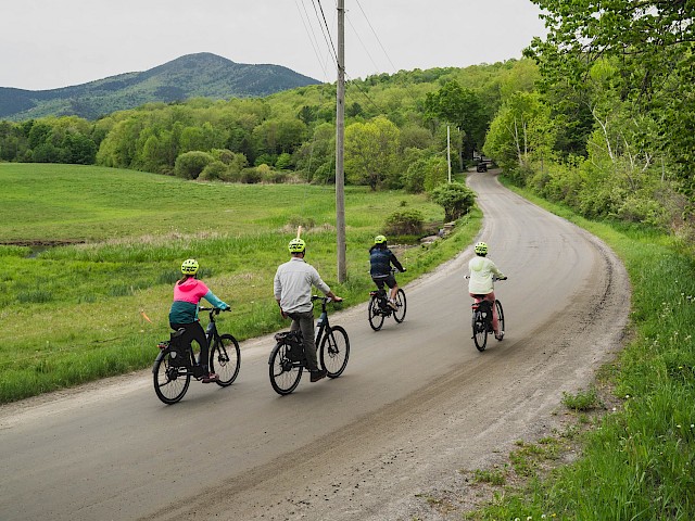 Four people are cycling on a curved dirt road through lush green countryside with mountains in the background.