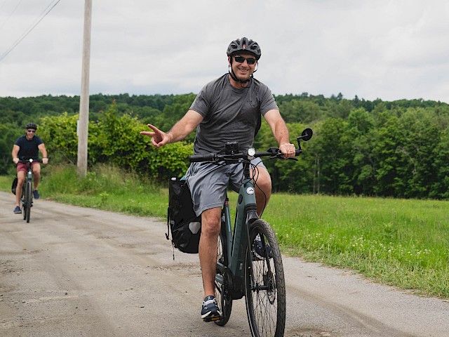 Two people ride bikes on a rural dirt road, smiling and making peace signs; the scenery is green and cloudy.