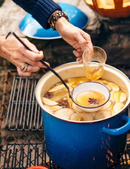 Someone is preparing mulled cider with apple slices, spices, and honey over an outdoor grill, likely for a cozy fall gathering.