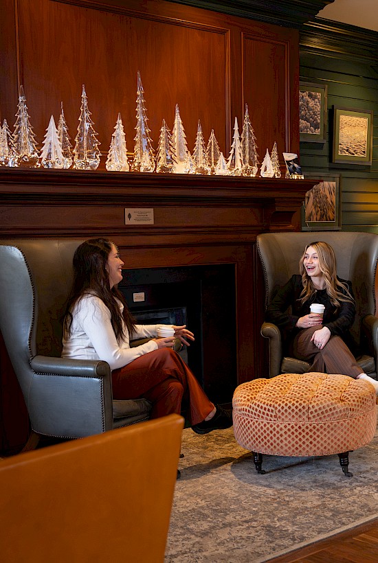 Four people are relaxing and chatting in a cozy, warmly-lit hotel lobby with comfortable chairs and festive decorations.